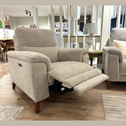The Delta Electric Recliner, upholstered in beige easy-clean fabric for modern comfort and practicality, is displayed in a showroom with wooden flooring. Positioned next to a matching armchair, side table, and decorative pillows, it features an extended footrest that combines style and relaxation.