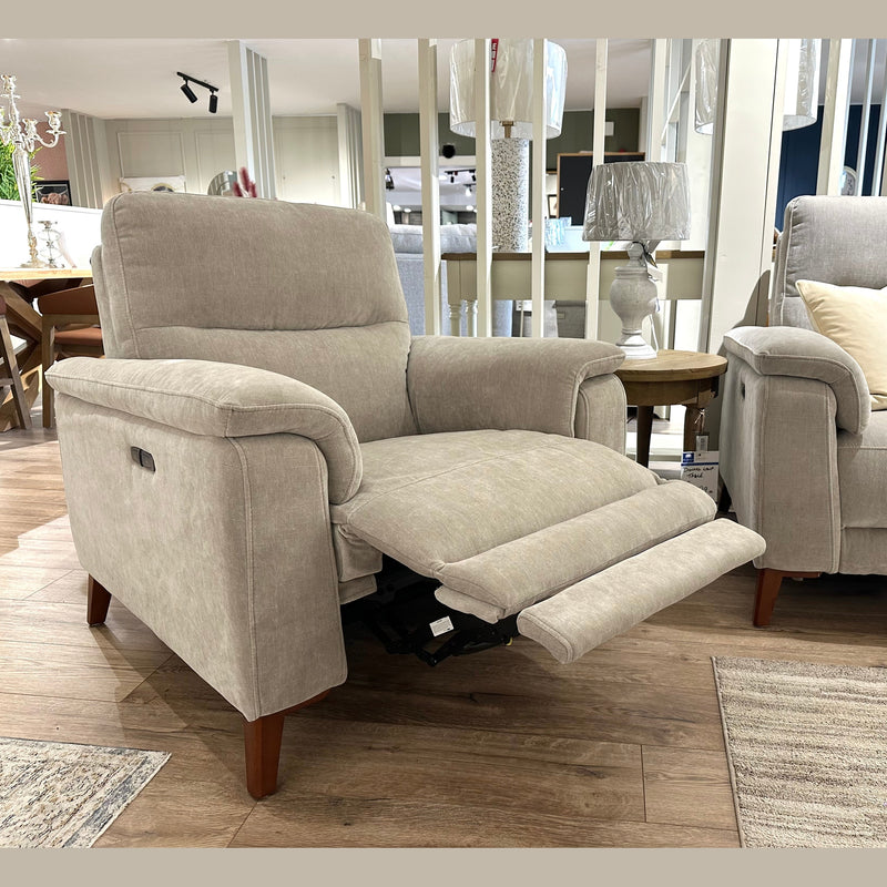 The Delta Electric Recliner, upholstered in beige easy-clean fabric for modern comfort and practicality, is displayed in a showroom with wooden flooring. Positioned next to a matching armchair, side table, and decorative pillows, it features an extended footrest that combines style and relaxation.