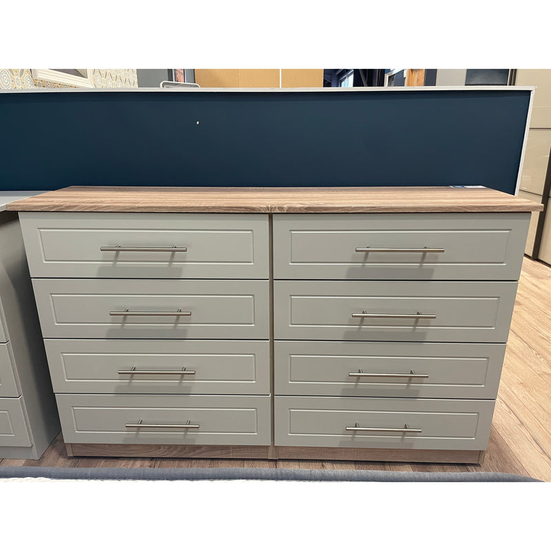 An image of a beige Millwood 8-drawer chest of drawers with a dark blue wall behind it, displayed in a showroom setting.