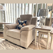 A Lorenzo Electric Recliner, upholstered in taupe washable fabric and featuring a beige finish with a floral cushion, sits beside a white lamp on a small table in a sunlit living room showroom with wooden floors and modern decor.