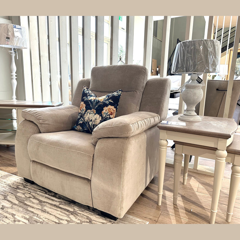 A Lorenzo Electric Recliner, upholstered in taupe washable fabric and featuring a beige finish with a floral cushion, sits beside a white lamp on a small table in a sunlit living room showroom with wooden floors and modern decor.