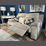 The Lorenzo Electric Recliner, upholstered in modern taupe washable fabric and accented with floral and geometric pillows, is featured in a cozy living room. In the background are two lamps, a white console table, mirrors, and blue walls.