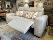 The Lennox Electric Recliner Sofa, upholstered in a modern light-gray fabric, is featured in a stylish living room setting with two patterned accent pillows. The left seat’s footrest is extended, showcasing the sofa’s luxurious electric recliner functionality. Complementary lamps, a rug, and additional contemporary furniture complete the scene.