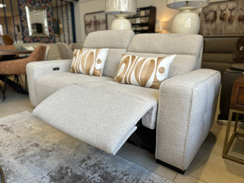 The Lennox Electric Recliner Sofa, upholstered in a modern light-gray fabric, is featured in a stylish living room setting with two patterned accent pillows. The left seat’s footrest is extended, showcasing the sofa’s luxurious electric recliner functionality. Complementary lamps, a rug, and additional contemporary furniture complete the scene.