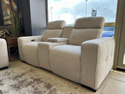 Two light gray Lennox Electric Recliner armchairs with a shared center console are displayed in a showroom near large windows. A beige rug is placed in front, with plants and other furniture visible in the background.