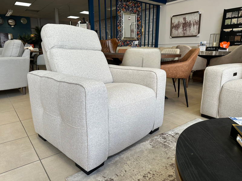 A Lennox Electric Recliner Sofa in light gray upholstery is showcased in a modern furniture showroom, surrounded by contemporary sofas, dining chairs, and tables. The space features neutral tones and stylish wall decor.