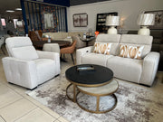 A Lennox Electric Recliner Sofa in beige, accompanied by a matching 2-seater sofa with patterned pillows and two round nesting coffee tables, is displayed on a rug in a modern furniture showroom. Additional luxury sofas and decor can be seen in the background.