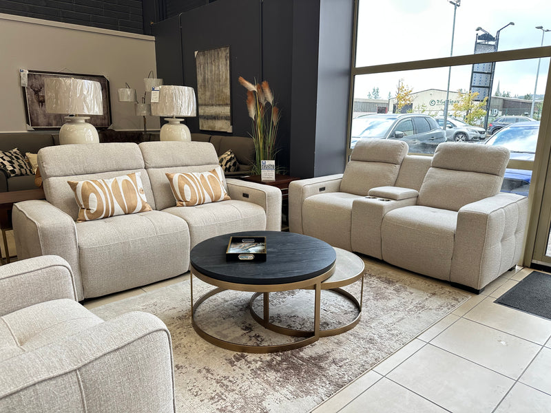 Modern furniture showroom featuring the luxurious Lennox Electric Recliner Sofa and a two seater sofa in beige, complemented by an armchair, round nested coffee tables, decorative pillows, a rug, stylish lamps, wall art, and large windows with an inviting outdoor view.