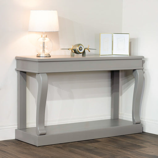 The Scroll Mink 130cm Console Table, featuring curved legs and a gray finish, holds a clear glass lamp, a small silver and gold airplane model, and two framed pictures. This entryway table stands against a white wall on a wood floor.