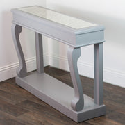 The Scroll Mink Antique Mirror Console, featuring a narrow, light gray design with elegantly curved legs and an antique mirror top, adds vintage charm to the space when placed against a white wall on dark wood flooring.