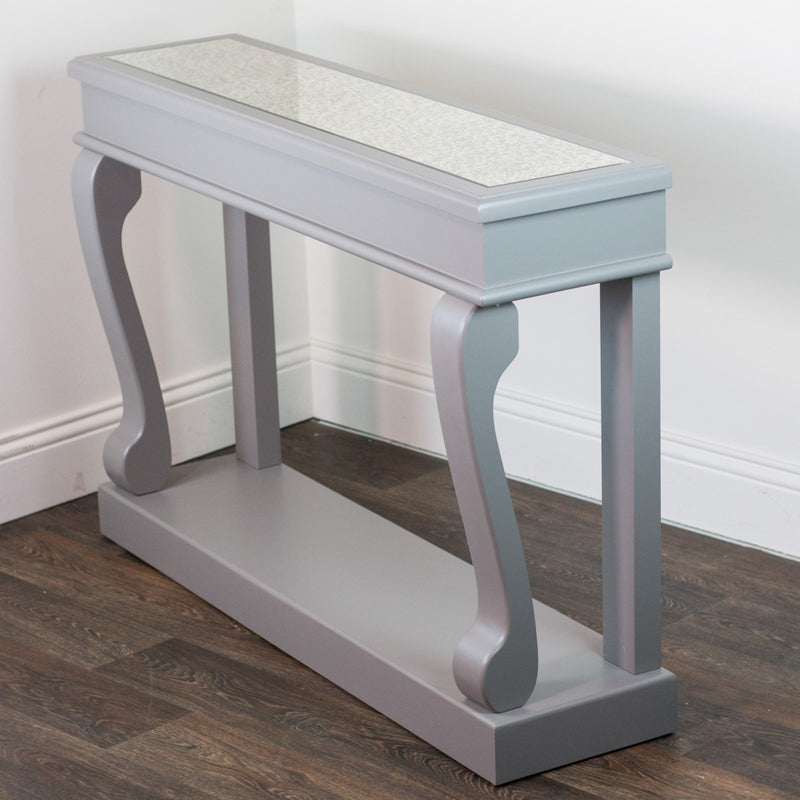 The Scroll Mink Antique Mirror Console, featuring a narrow, light gray design with elegantly curved legs and an antique mirror top, adds vintage charm to the space when placed against a white wall on dark wood flooring.