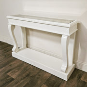 A Scroll Ivory Antique Mirror Console with curved legs sits against a white wall on a dark wood floor, featuring an elegant mirrored tabletop.