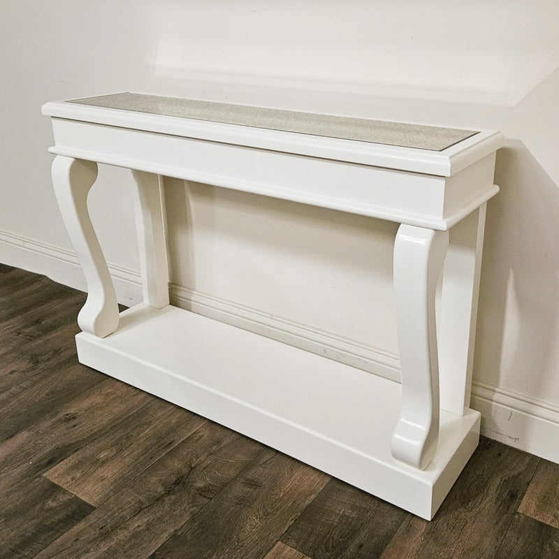 A Scroll Ivory Antique Mirror Console with curved legs sits against a white wall on a dark wood floor, featuring an elegant mirrored tabletop.