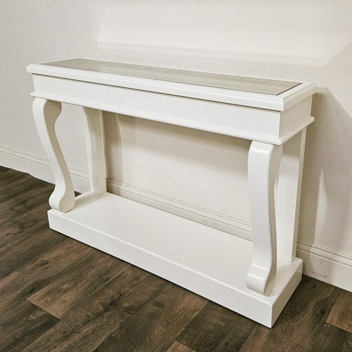 The Scroll Ivory Antique Mirror 120cm Console, featuring a white vintage finish with elegant curved legs and a lower shelf, is placed against a white wall on a dark wood floor.