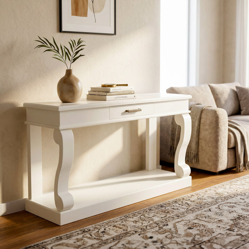 The Scroll 150cm Ivory Console, featuring curved, scroll-inspired legs and a convenient drawer, stands against a neutral wall near a beige couch and patterned rug in a sunlit living room. Topped with stacked books and a vase of green branches, it adds elegant functionality to the entryway.
