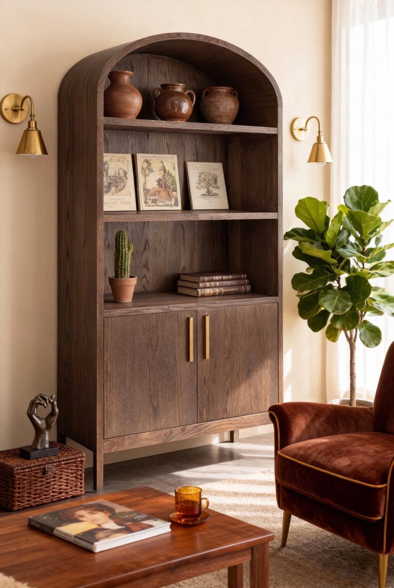 A cozy living room featuring the Sienna Tall Cabinet, a wooden bookshelf that doubles as practical living room storage, holding pots, books, and a cactus. A plant, vintage armchair, table with a cup, and decorative items complete the warm, sunlit space.