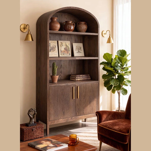 A cozy living room featuring the Sienna Tall Cabinet, a wooden bookshelf that doubles as practical living room storage, holding pots, books, and a cactus. A plant, vintage armchair, table with a cup, and decorative items complete the warm, sunlit space.