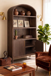 The Sienna Tall Cabinet functions as a bookshelf, displaying pottery, books, and a cactus against a beige wall accented with brass sconces. Nearby, a brown armchair sits beside a glass mug and potted plant in the sunlit, cozy room.