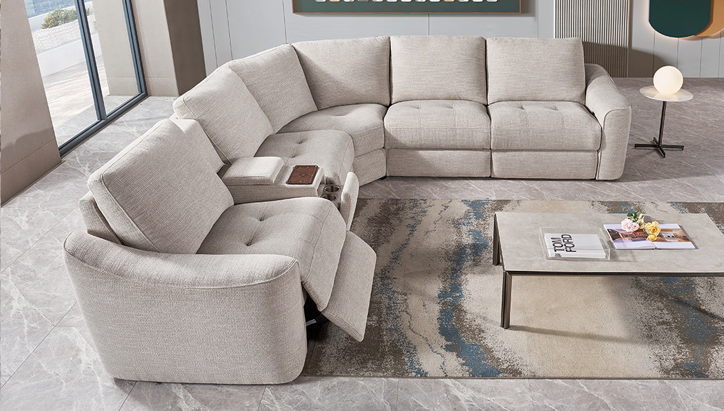 A modern living room features the Velora Corner Group modular sectional in light gray with an electric recliner, paired with a marble coffee table displaying books and decor atop a patterned rug on tiled flooring.