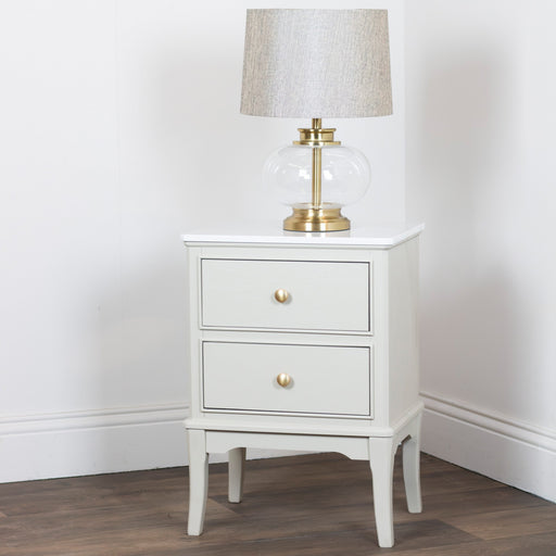 A Monroe 2 Drawer Locker in Stone with a marble top and gold knobs stands on wooden flooring, topped with a glass and brass table lamp with a beige fabric shade, set against a plain white wall.