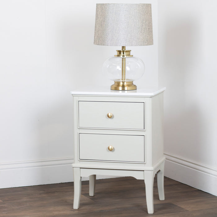 A Monroe 2 Drawer Locker in Stone with a marble top and gold knobs stands on wooden flooring, topped with a glass and brass table lamp with a beige fabric shade, set against a plain white wall.