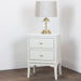 A Monroe 2 Drawer Locker in Stone with a marble top and gold knobs stands on wooden flooring, topped with a glass and brass table lamp with a beige fabric shade, set against a plain white wall.