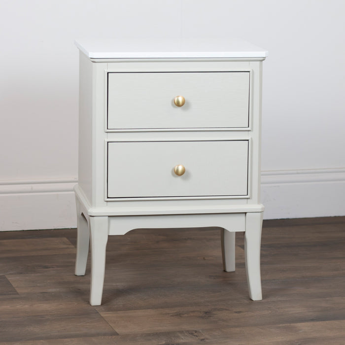 The Monroe 2 Drawer Locker - Stone, a compact white wood accent table with a marble top and round gold drawer knobs, stands on a wood floor against a plain white wall.