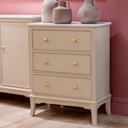 The Monroe Large 3 Drawer Locker - Stone stands on wooden flooring against a pink wall, featuring yellow knobs and a marble top adorned with two decorative ceramic vases, bringing elegance to the space.