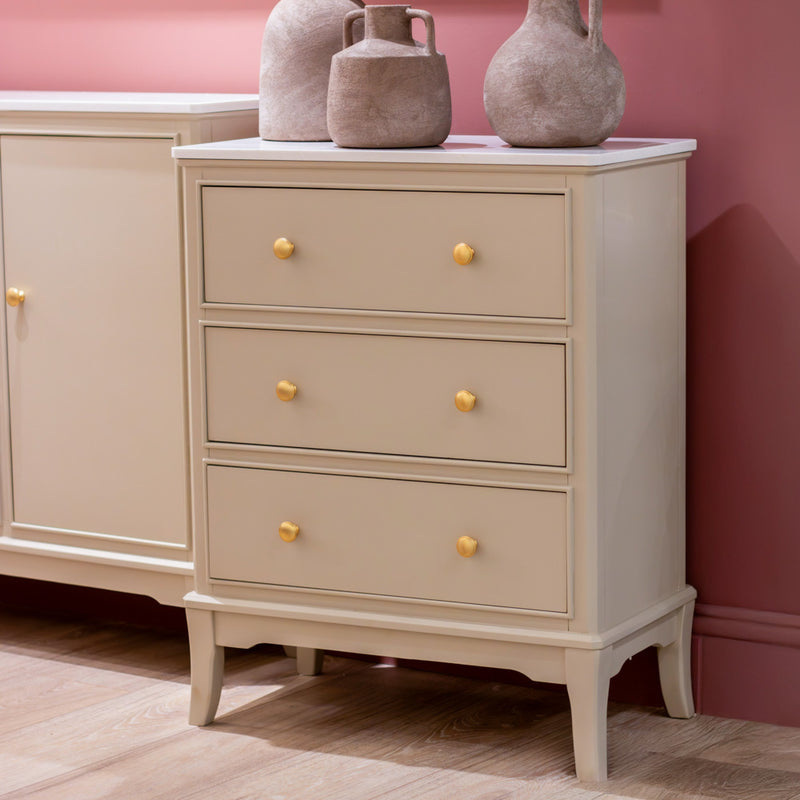 The Monroe Large 3 Drawer Locker - Stone stands on wooden flooring against a pink wall, featuring yellow knobs and a marble top adorned with two decorative ceramic vases, bringing elegance to the space.