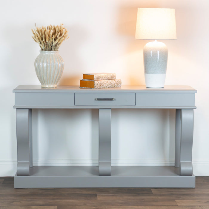 The Large Scroll Console with Drawer in warm grey features a center drawer and displays two books, a large vase of dried wheat, and a white lamp. It stands against a white wall on a wood floor.