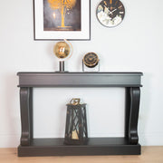 The Scroll Console - Antique Mirror Top displays a gold globe and clock, with a lantern beneath. Above, framed palm art and a floral clock adorn the white wall, blending minimalist style with vintage charm.