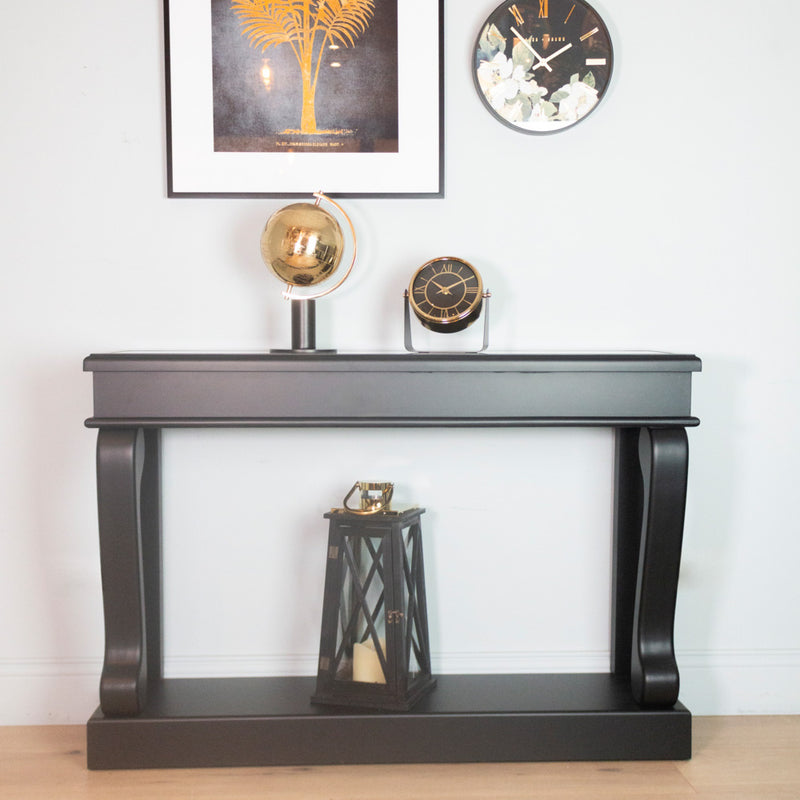 The Scroll Console - Antique Mirror Top displays a gold globe and clock, with a lantern beneath. Above, framed palm art and a floral clock adorn the white wall, blending minimalist style with vintage charm.