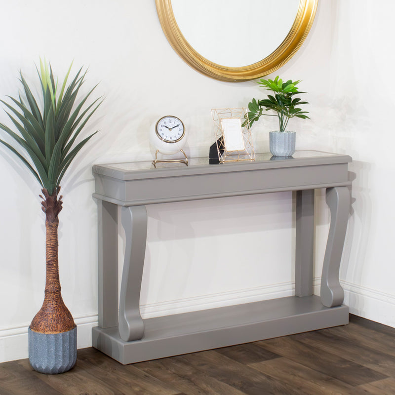 A Scroll Console - Antique Mirror Top stands by a white wall, topped with a clock, photo frame, and potted plants for vintage charm. Above hangs a round gold mirror, while a tall floor plant accents the dark wood floor alongside.