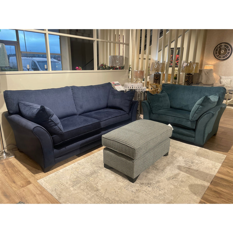 A living room display featuring a modern curved arm sofa in navy blue, the Savoy Armchair in teal, and a patterned ottoman on a light rug, set against wooden floors and decorative accents—highlighting Irish-made furniture.