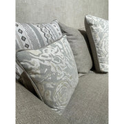 A close-up of the Shelly Angle Seatee, a grey fabric sofa made in Ireland, featuring three decorative pillows—floral, geometric, and solid grey—set against a textured light grey wall.