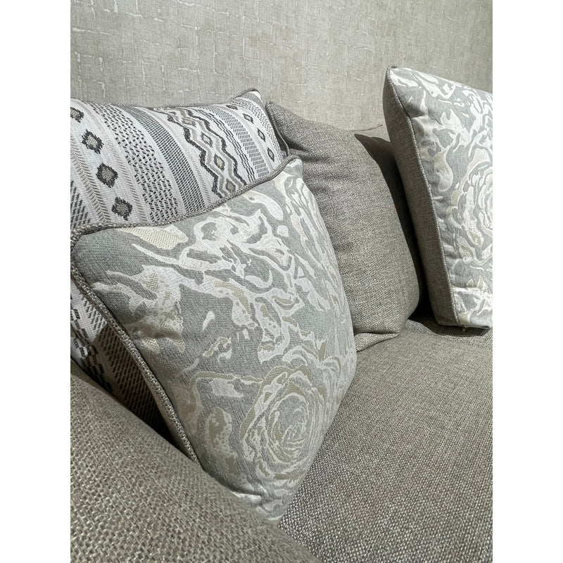 A close-up of the Shelly Angle Seatee, a grey fabric sofa made in Ireland, featuring three decorative pillows—floral, geometric, and solid grey—set against a textured light grey wall.