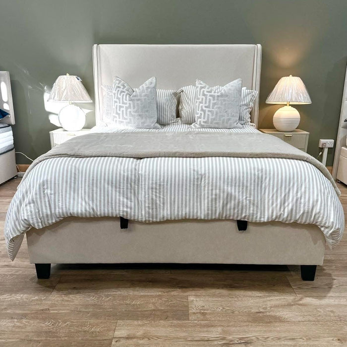 A modern bedroom with the Rivera Storage Bedframe in beige linen, gray and white striped bedding, patterned pillows, two white bedside tables with matching lamps, set against a muted green wall and wood floor.