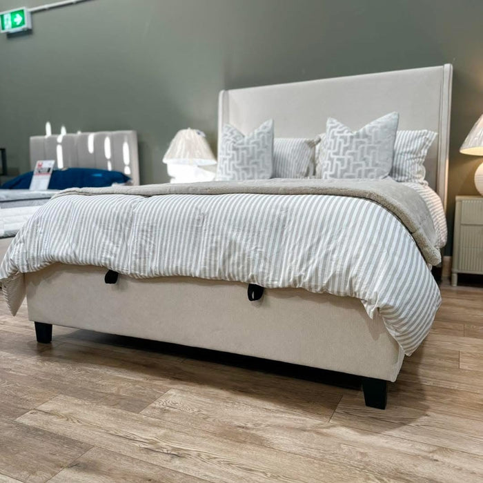 A neatly made Rivera Storage Bedframe with a light-toned upholstered headboard, striped bedding, and decorative pillows is displayed in a showroom featuring wooden floors and bedside lamps.