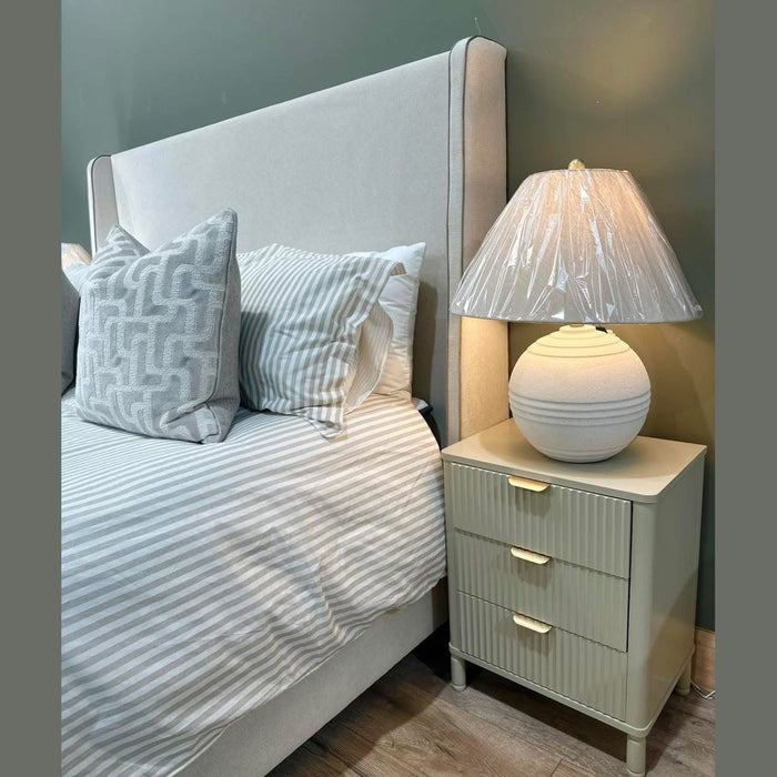 A Rivera Storage Bedframe with striped bedding and two large pillows stands next to a beige three-drawer nightstand topped with a round white pleated-shade lamp still wrapped in plastic.