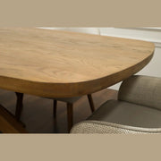 A close-up of the Ashton Dining Table in light solid ash wood with rounded edges, paired with an upholstered chair in patterned fabric and a cushioned seat, set against a white wall.