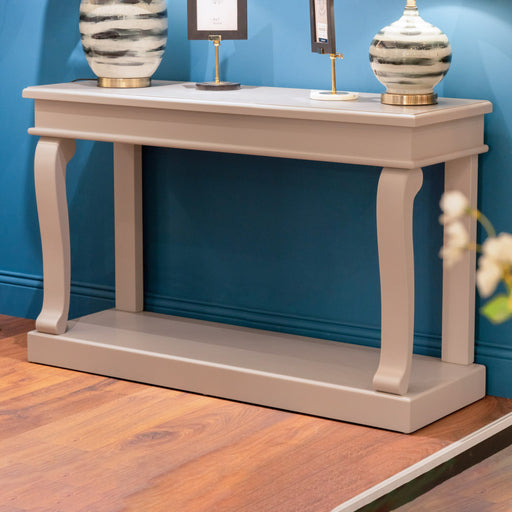 The Scroll console 130cm in light gray, featuring curved legs, stands against a blue wall on a wooden floor. Patterned vases and framed photos decorate the tabletop.