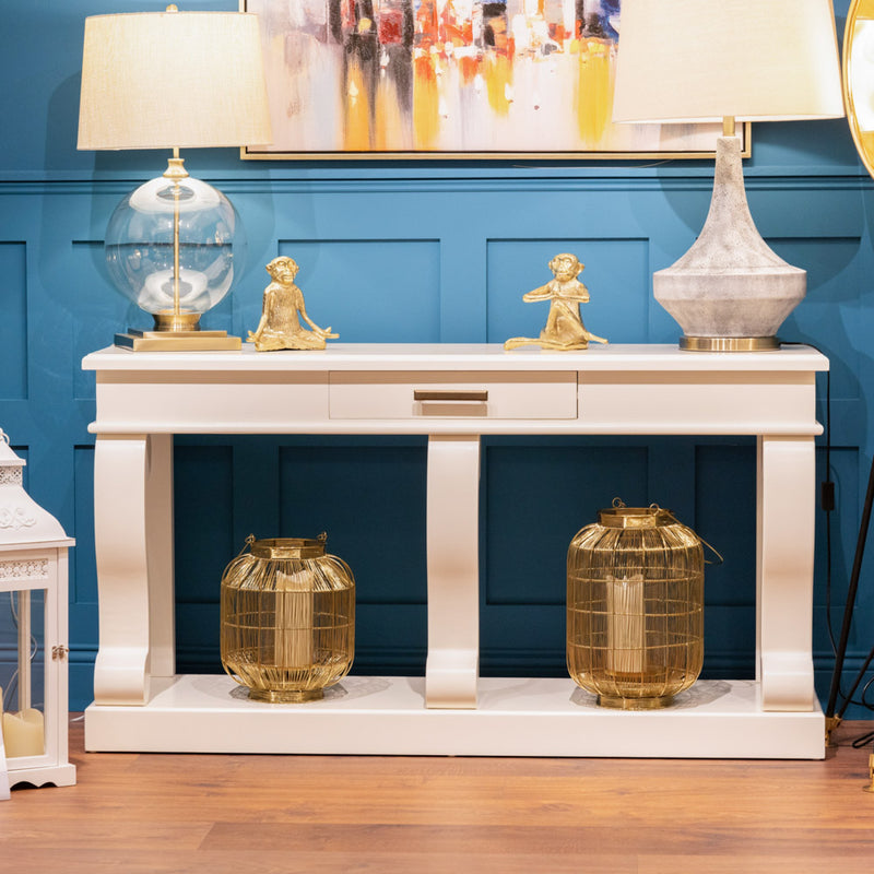 A Large scroll console with drawer is styled with two gold lanterns on the bottom shelf, two gold Buddha figurines and two lamps on top, set against a blue paneled wall and wooden floor.