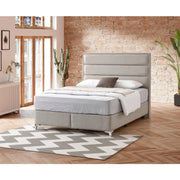 A gray Iris Storage Bedframe with chrome feet, placed in a room with a decorative plant, a stylish rug, and a brick wall in the background.