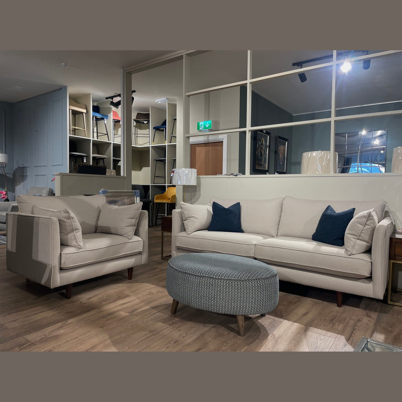 A modern living room featuring the Hugo Irish-made contemporary sofa with dark blue Aquaclean pillows, a beige armchair, round gray ottoman, and wooden flooring. Shelves and chairs are visible in the background.