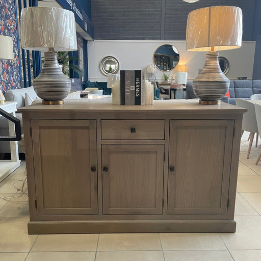 A Marlo Sideboard in American oak, topped with two lamps and three books, stands in a modern showroom featuring mirrors, tables, chairs, and hanging white paper lanterns in the background.