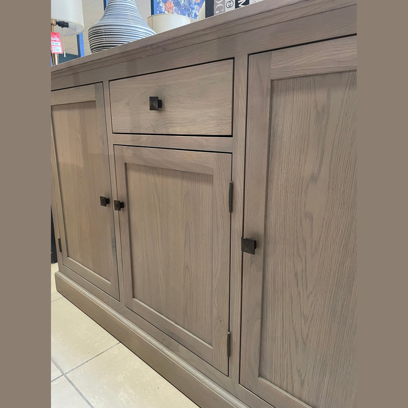 A close-up of the Marlo Sideboard in light wood, featuring three black square handles on two cabinet doors and a drawer, stands on a tiled floor with decorative vases displayed on top.