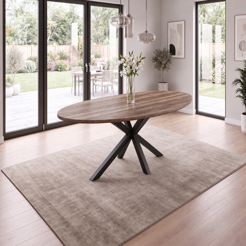 Elson Oval Table with a black metal star base, holding a glass vase of white flowers, centered on an area rug in a sunlit room.