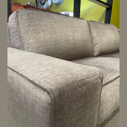 A close-up of the Sqaured Sofabed, a modern light brown fabric sofa with wide armrests, shown indoors against a yellow and black wall.