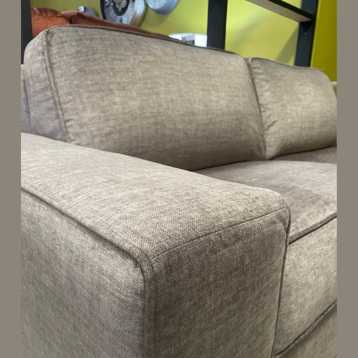 A close-up of the Sqaured Sofabed, a modern light brown fabric sofa with wide armrests, shown indoors against a yellow and black wall.