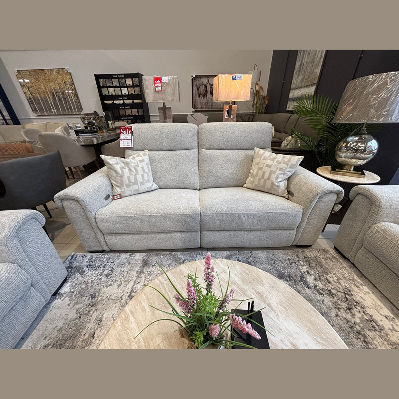 The Luna Electric Recliner, upholstered in light gray fabric and accented with two patterned cushions, is showcased in a showroom with a Sale tag on the left armrest and a coffee table featuring a pink floral arrangement in front.